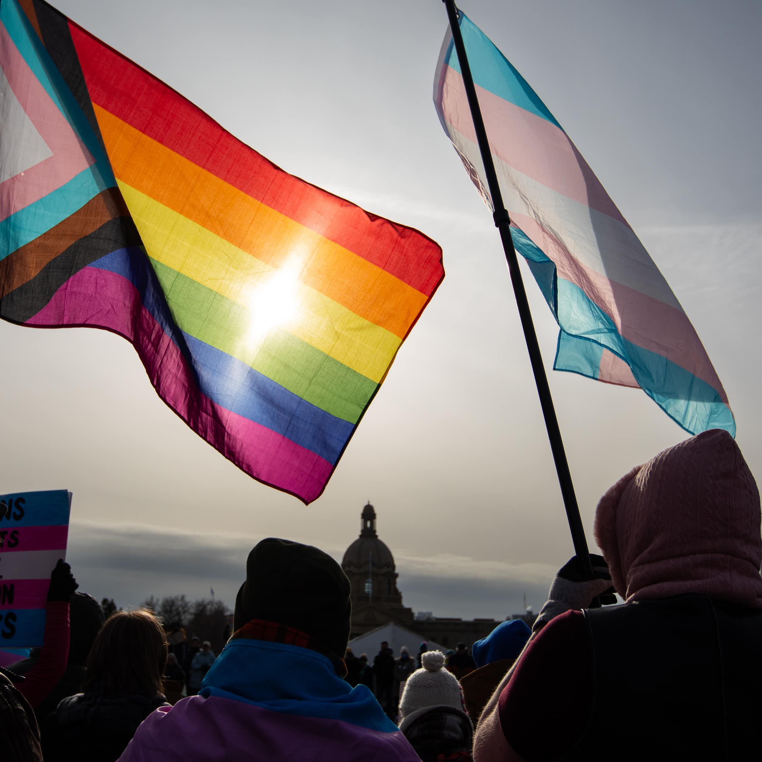 A rainbow LGBTQ+ flag and a trans flag seen with a legislative building in the background.