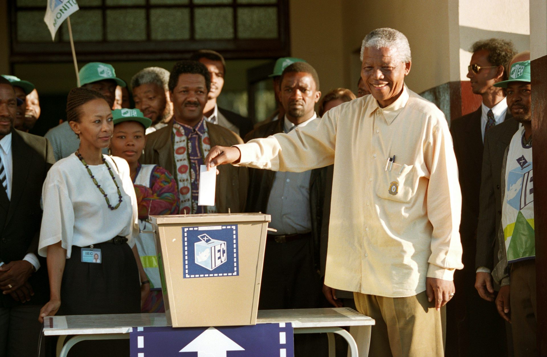 A grey-haired and smiling man wearing a loose long-sleeved shirt holds a piece of paper above a ballot box, while other people stand around him