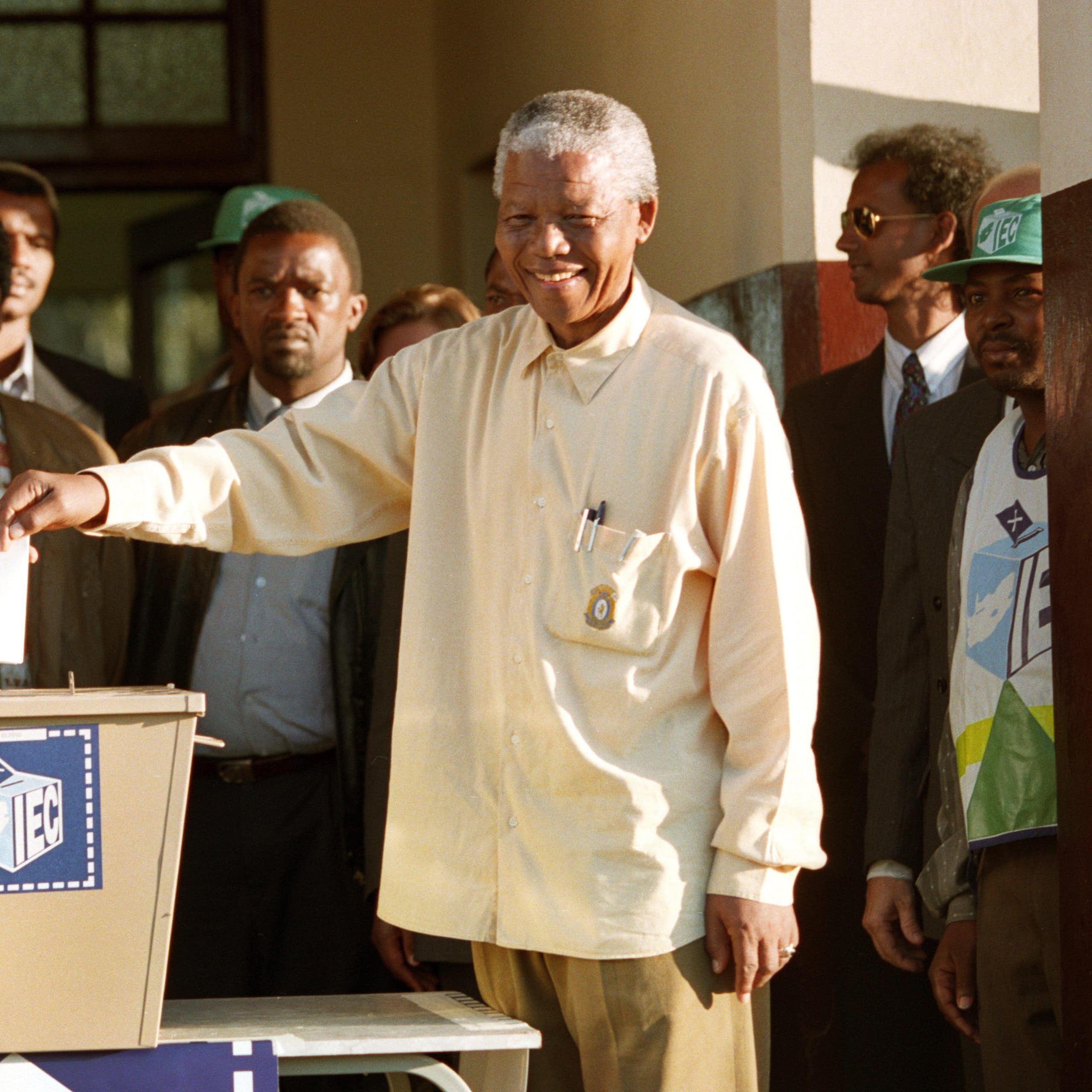 A grey-haired and smiling man wearing a loose long-sleeved shirt holds a piece of paper above a ballot box, while other people stand around him