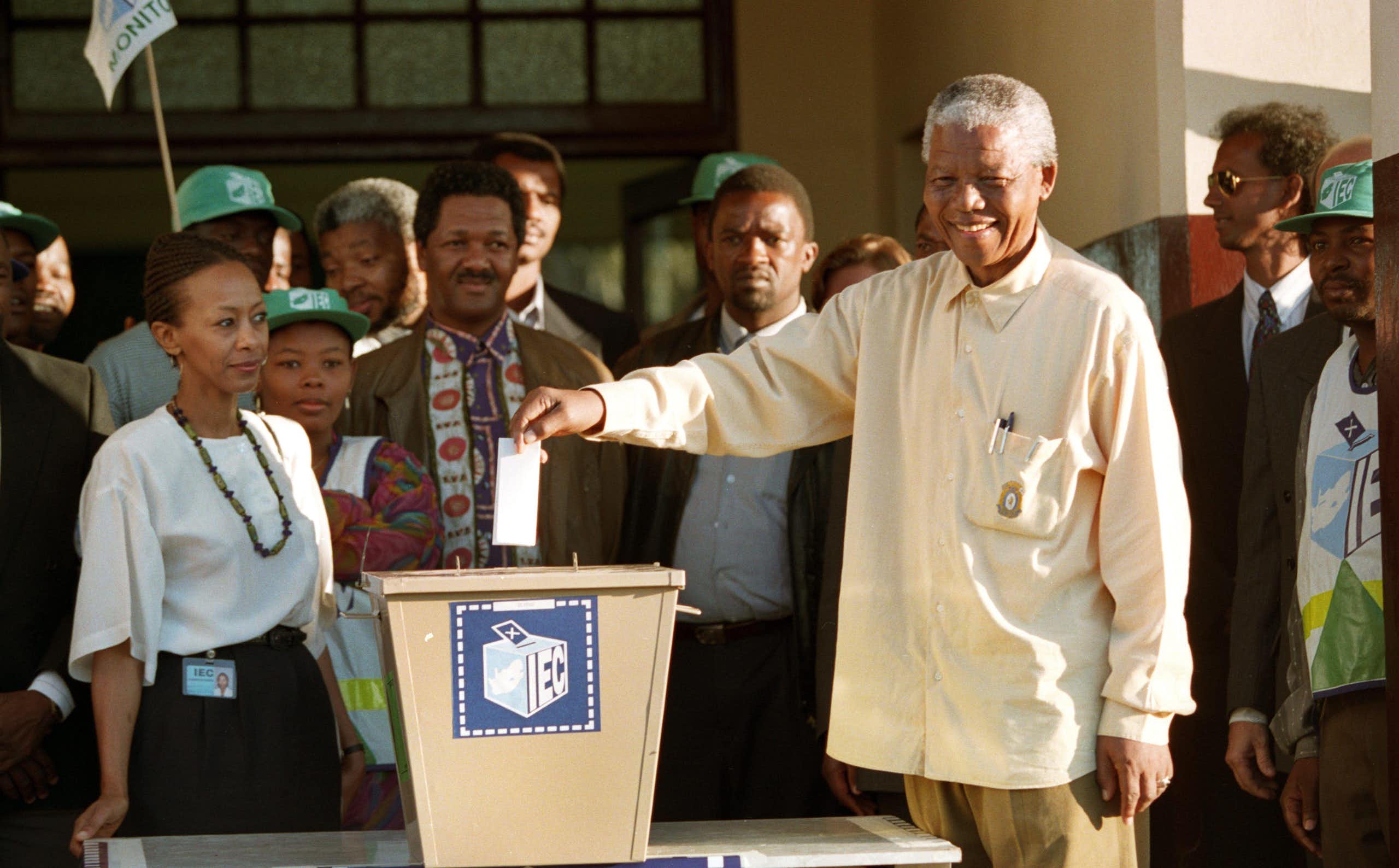 A grey-haired and smiling man wearing a loose long-sleeved shirt holds a piece of paper above a ballot box, while other people stand around him