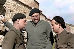 Two men dressed as soldiers talk face to face with a third man in the middle, wearing a beret.