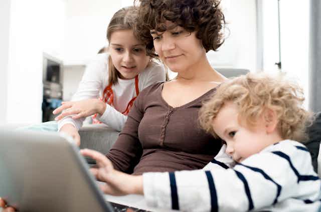 A woman with brown curly hair sits with two children looking at a laptop.