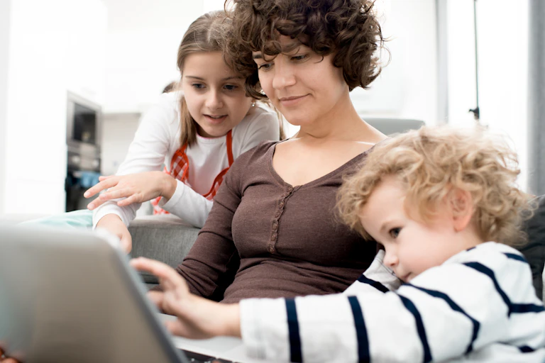 A woman with brown curly hair sits with two children looking at a laptop.