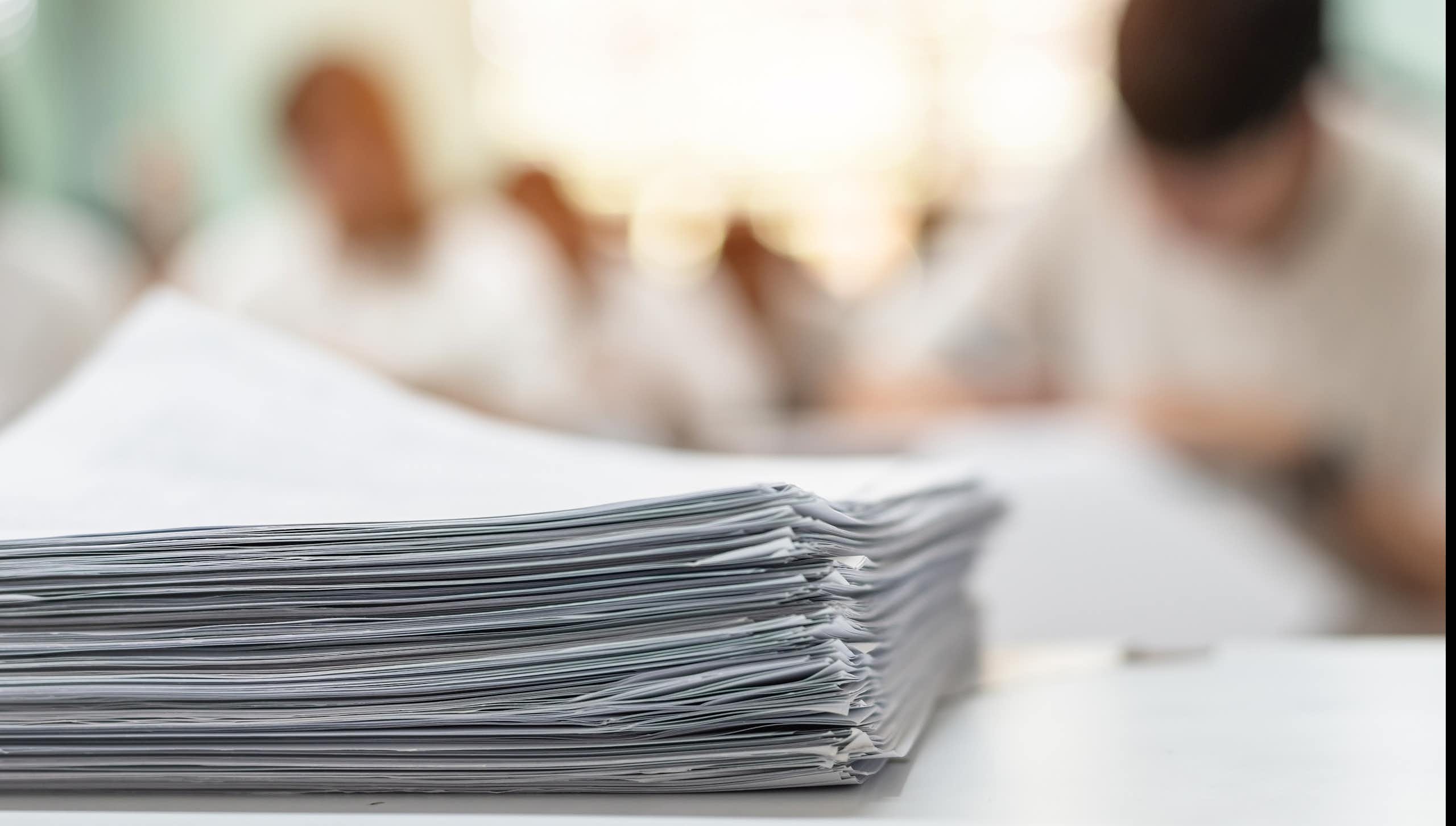 A huge pile of paperwork in the foreground with the blurred out background shows people working at desks.