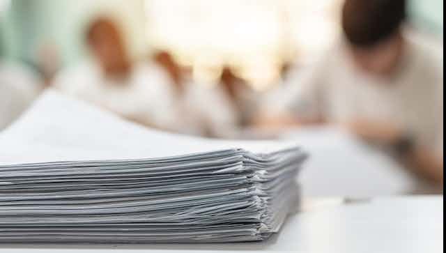 A huge pile of paperwork in the foreground with the blurred out background shows people working at desks.