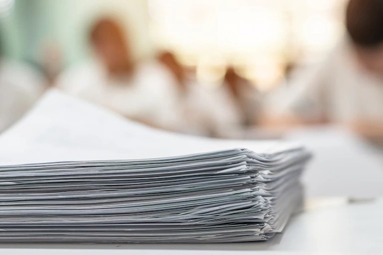 A huge pile of paperwork in the foreground with the blurred out background shows people working at desks.