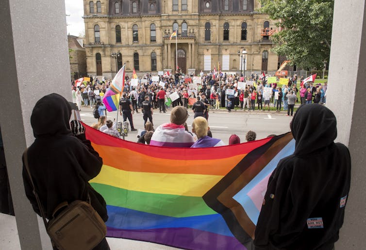 A rainbow and pink and grey triangle flag seen extended between two people and across the street are other people holding signs.
