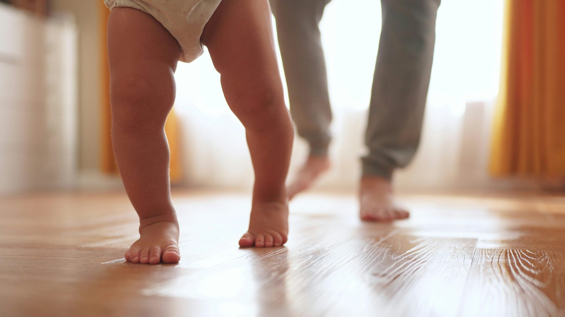a baby's legs as they walk with a parent behind them