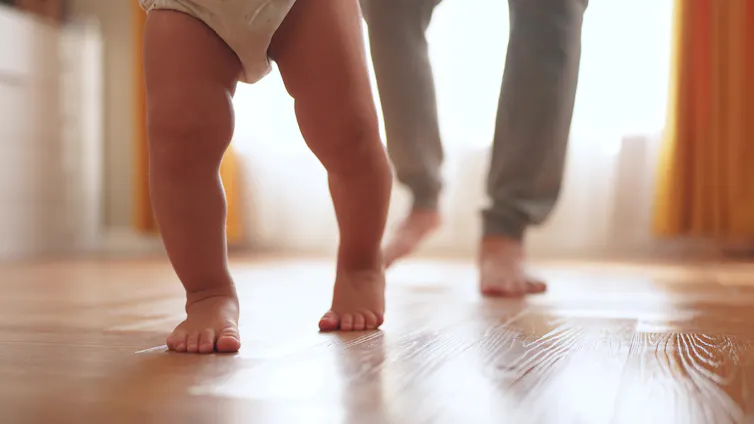 a baby's legs as they walk with a parent behind them