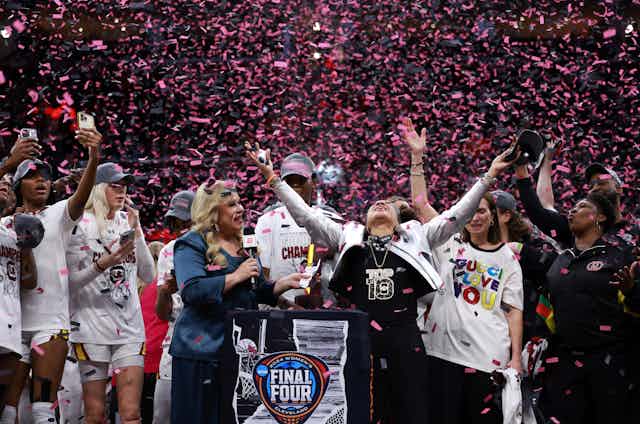 Pink confetti falls from the rafters of a basketball arena as a female coach and her players celebrate.