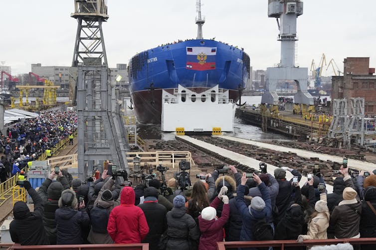 A large blue boat at a shipyard filled with a crowd of people.