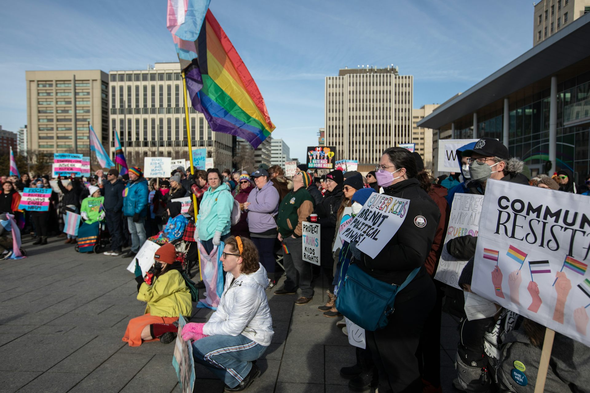 People standing in a semi-circle holding banners, one has a rainbow flag.