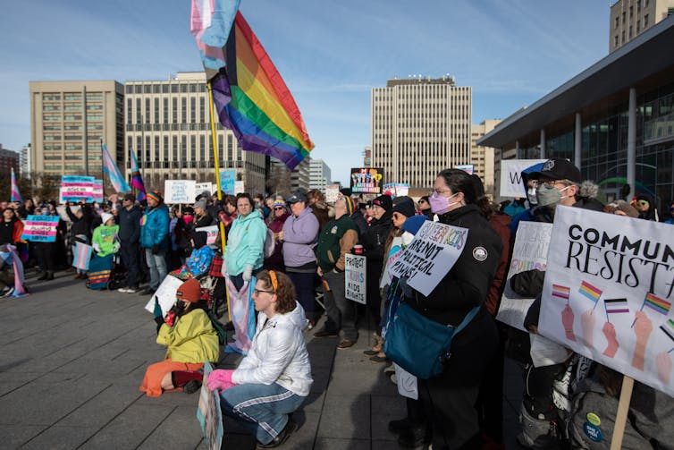 People standing in a semi-circle holding banners, one has a rainbow flag.