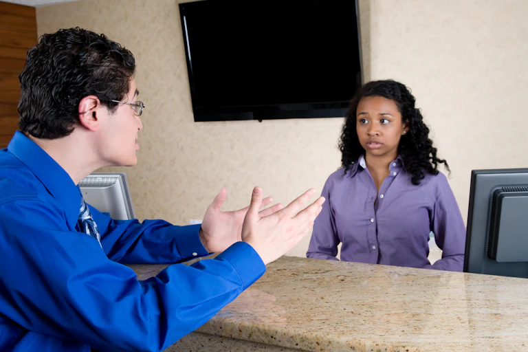 An angry male customer yells at a female employee staffing the check-in desk of a hotel.