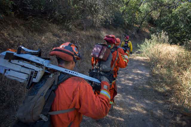 Inmate firefighters in orange and carrying equipment for fighting fires follow a crew chief along a path.
