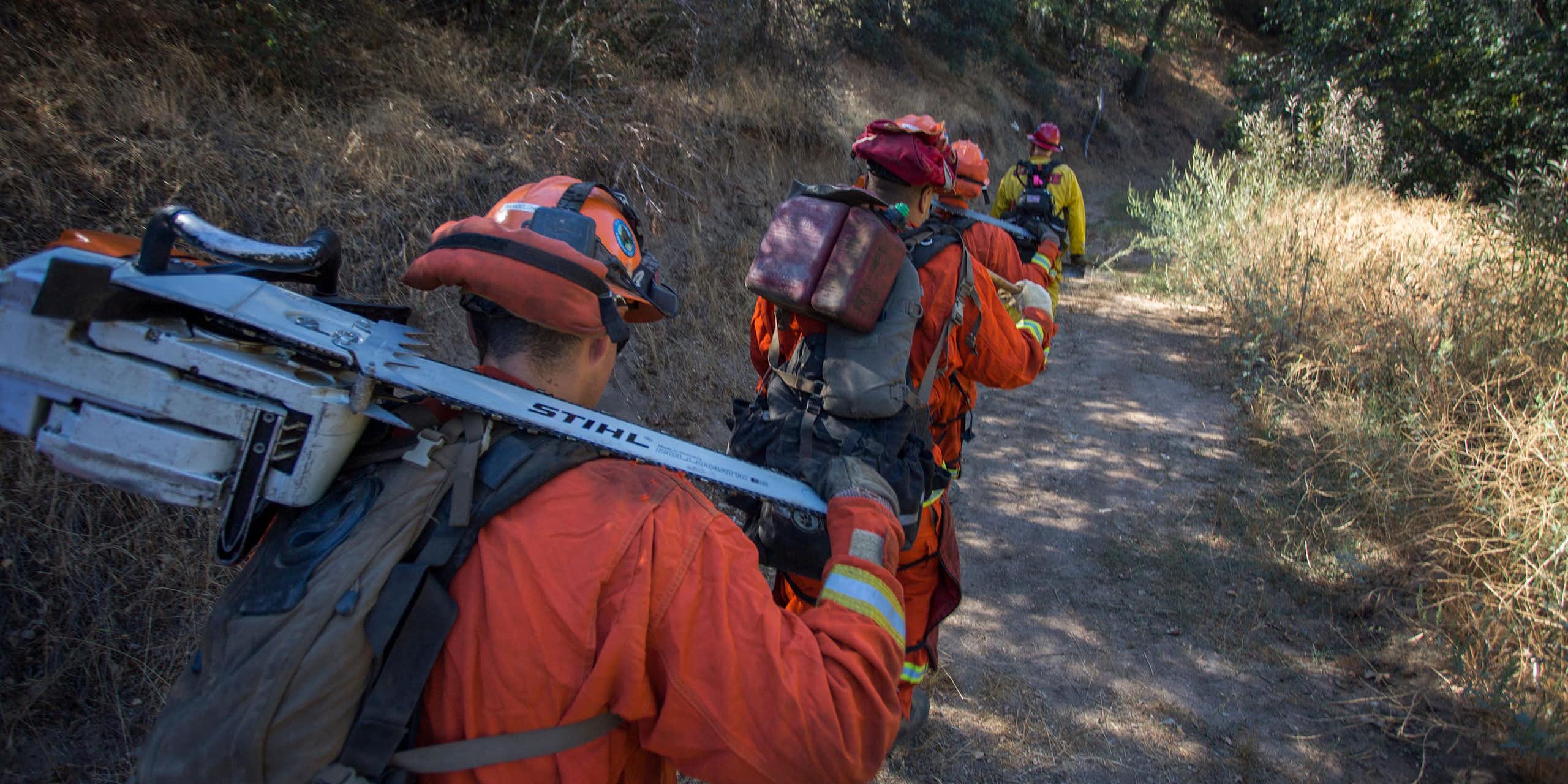 Inmate firefighters in orange and carrying equipment for fighting fires follow a crew chief along a path.