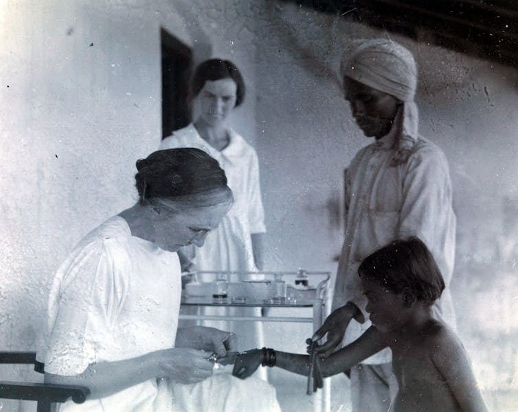 A black and white photo of a woman poking a needle into a child's wrist, with two other women in the background watching.