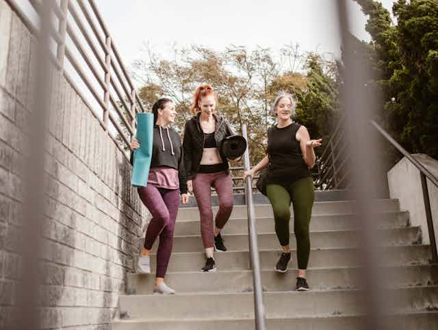 three women walk down steps carrying yoga mats, dressed for exercise
