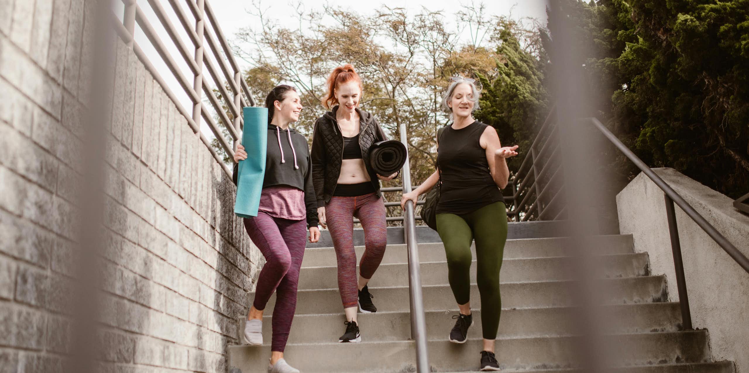 three women walk down steps carrying yoga mats, dressed for exercise
