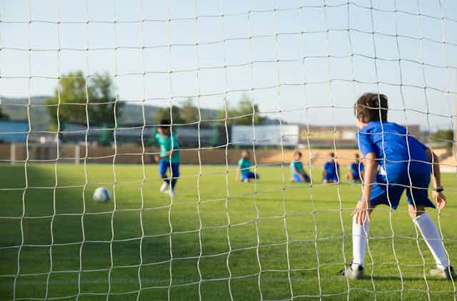 A boy prepares to kick a ball into a soccer net. Another boy waits to stop it. 