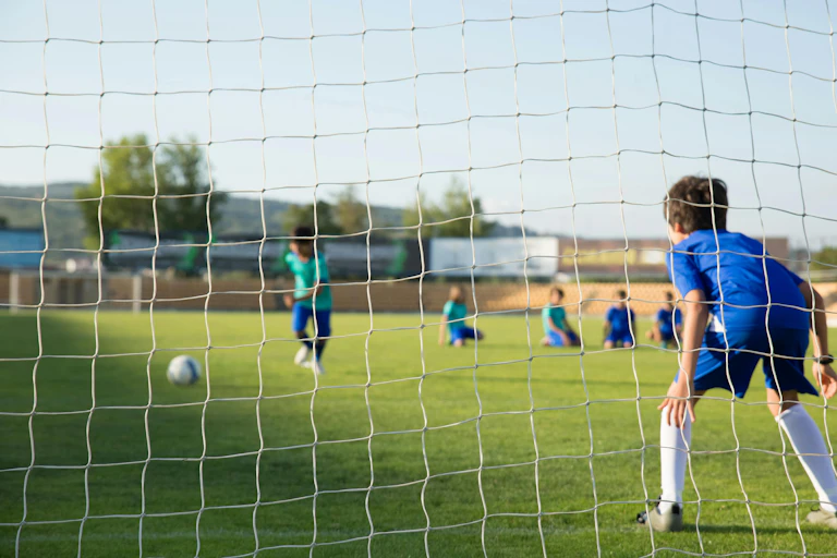 A boy prepares to kick a ball into a soccer net. Another boy waits to stop it.