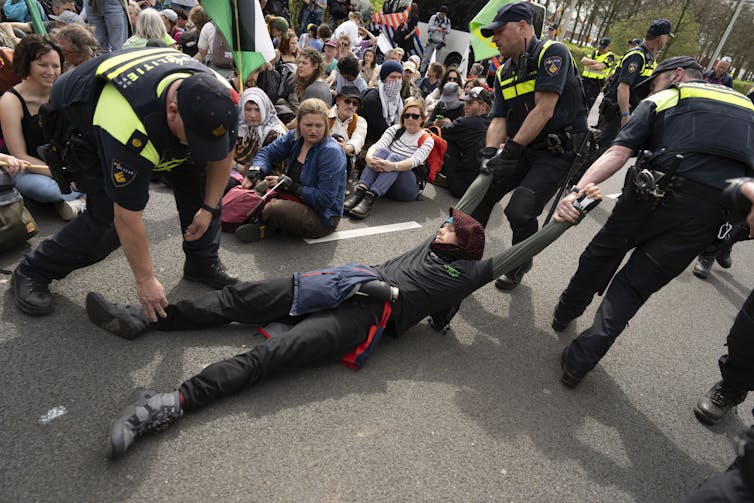A man at a protest being dragged by police. Other protestors sit on the ground in the background.