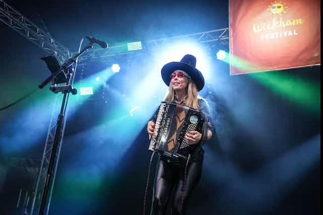 A woman in a tall black hat plays an accordion on stage.