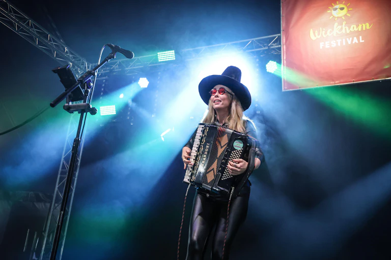 A woman in a tall black hat plays an accordion on stage.