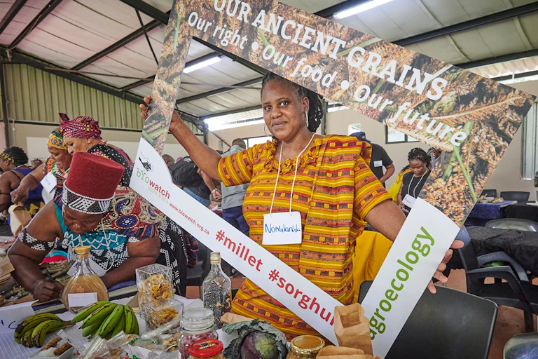 A woman selling vegetables and grains holds up a sign saying 'ancient grains: our right, our food, our future'