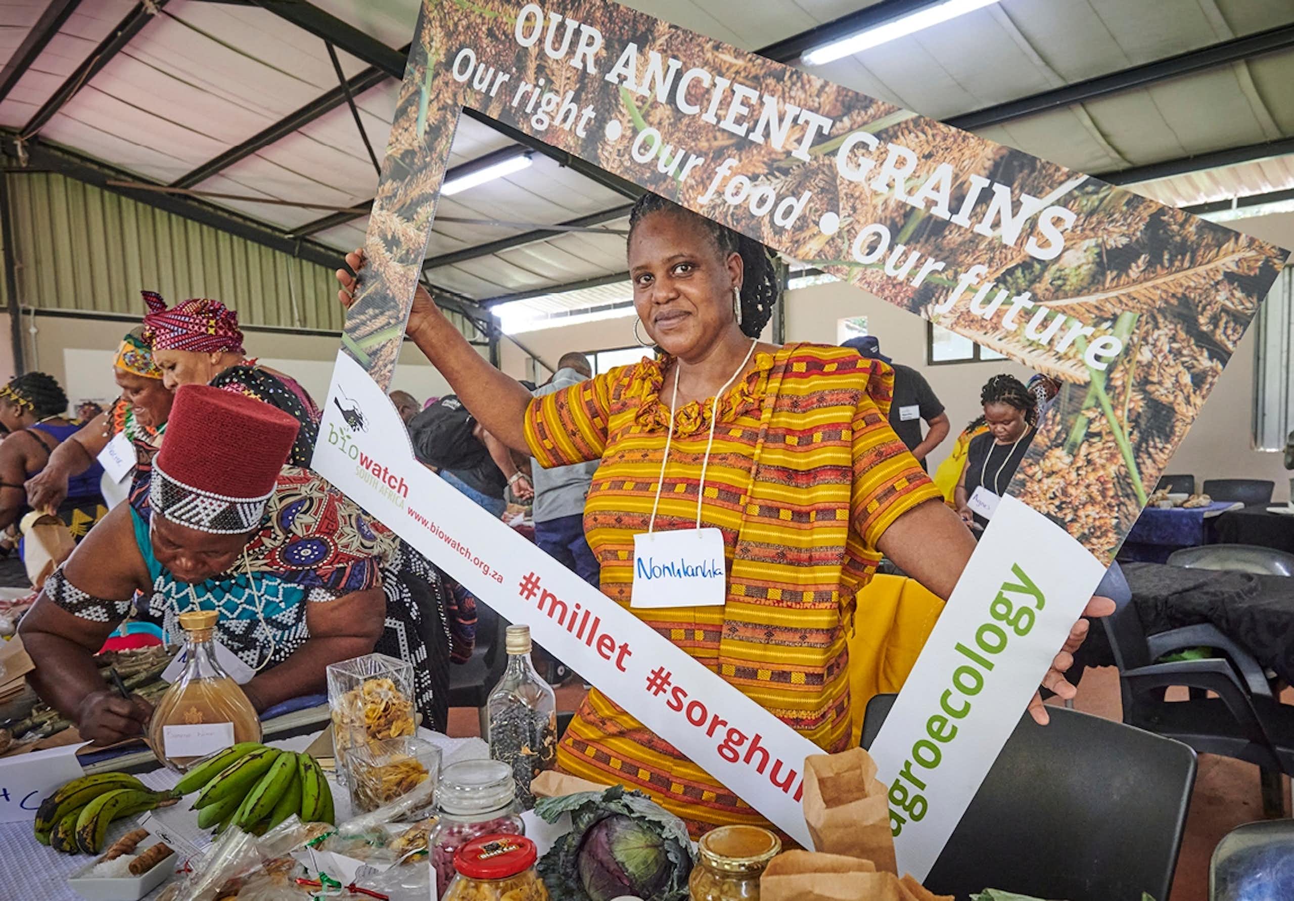 A woman selling vegetables and grains holds up a sign saying 'ancient grains: our right, our food, our future'