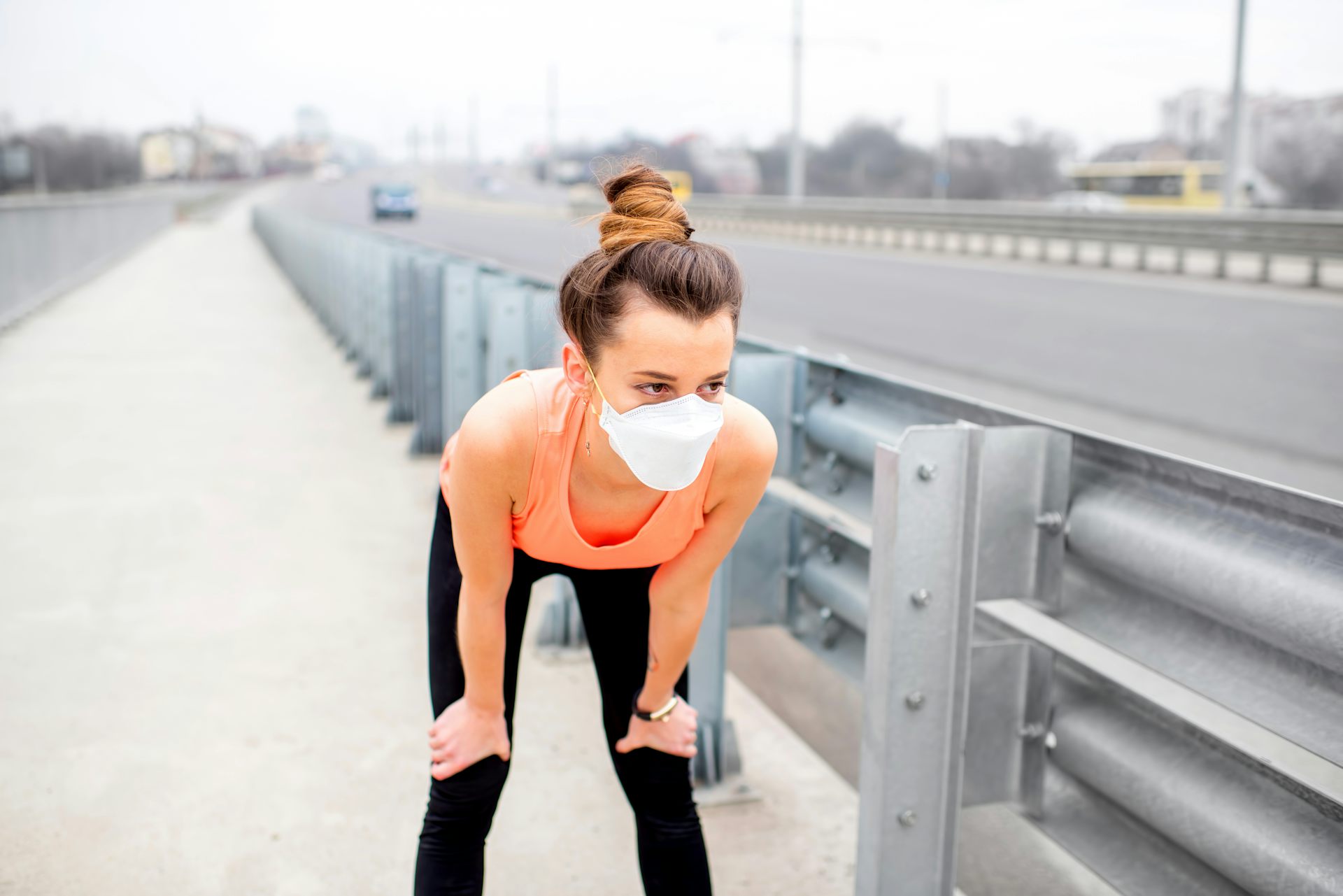 While jogging through town, a young woman rests with her hands on her knees and a protective mask over her face.