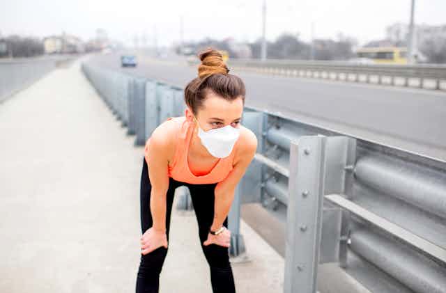 While jogging through town, a young woman rests with her hands on her knees and a protective mask over her face.
