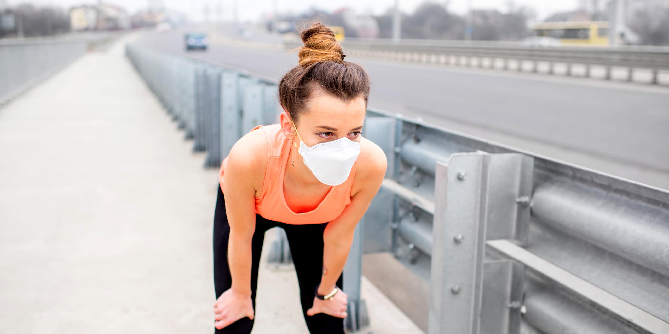 While jogging through town, a young woman rests with her hands on her knees and a protective mask over her face.