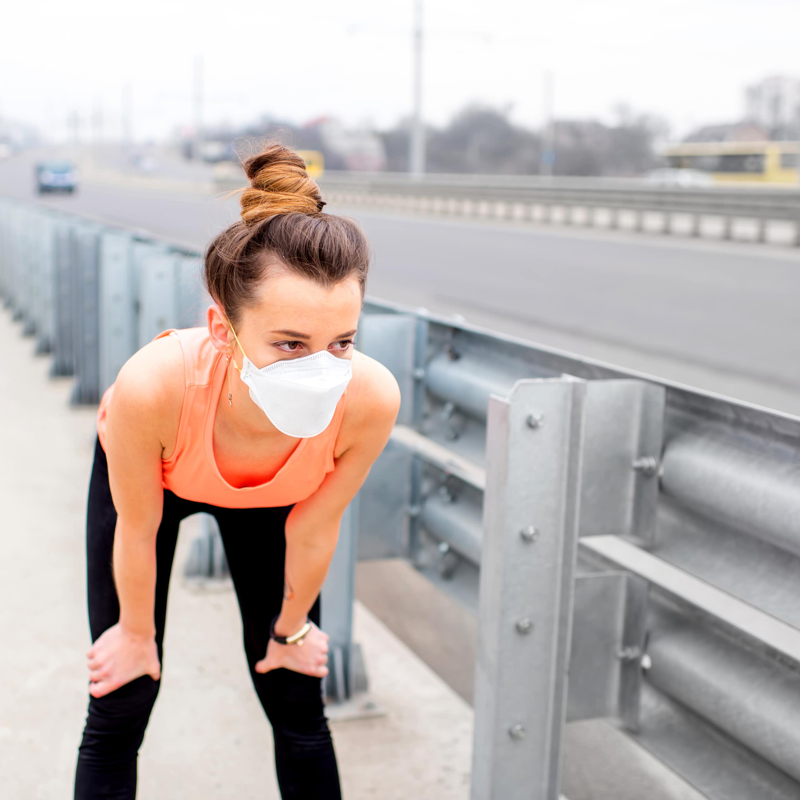 While jogging through town, a young woman rests with her hands on her knees and a protective mask over her face.