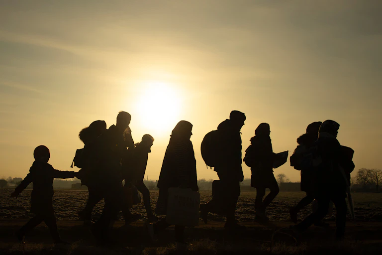 Side view of silhouettes of a group of people of different ages and genders walking across a field with the sun illuminating the background
