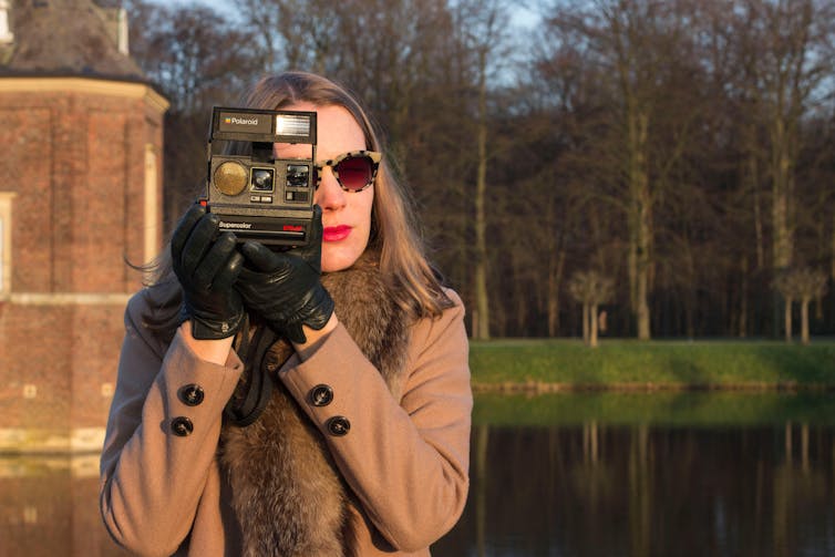 Woman wearing sunglasses holds a Polaroid camera to her eye