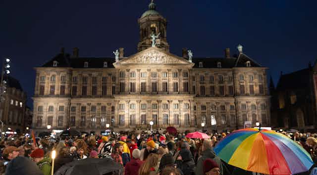Thousands of people outside a lit palace at night, one carrying a rainbow umbrella.