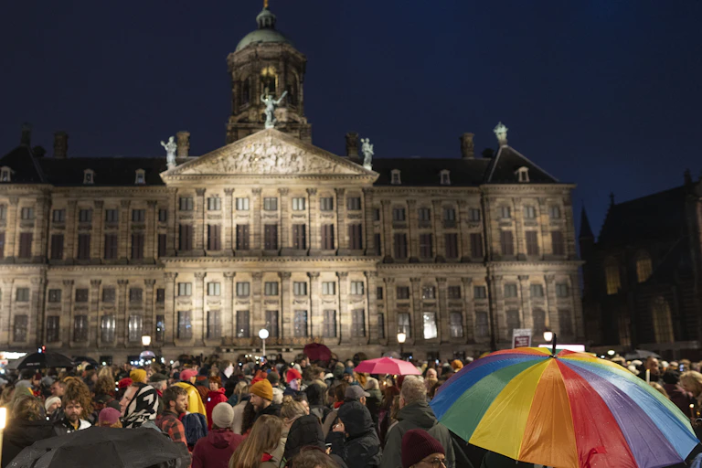 Thousands of people outside a lit palace at night, one carrying a rainbow umbrella.