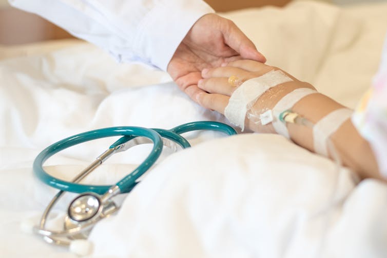 Child's hand with IV placed in wrist holding hand of person wearing white coat, both hovering over a stethoscope on a bed