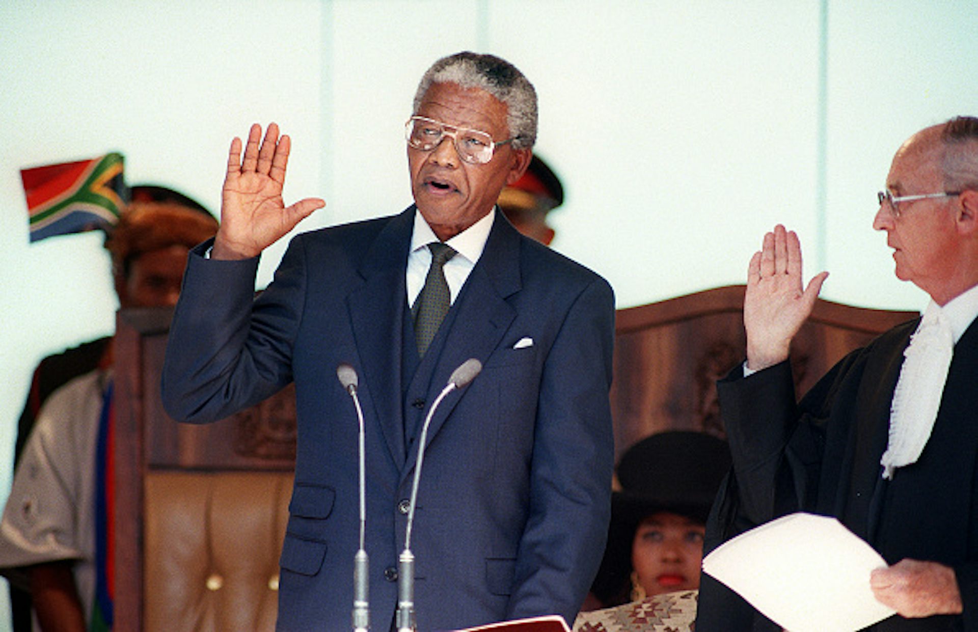A man wearing a suit and tie raises his right hand to take an oath, wtacxhed by a male judge.