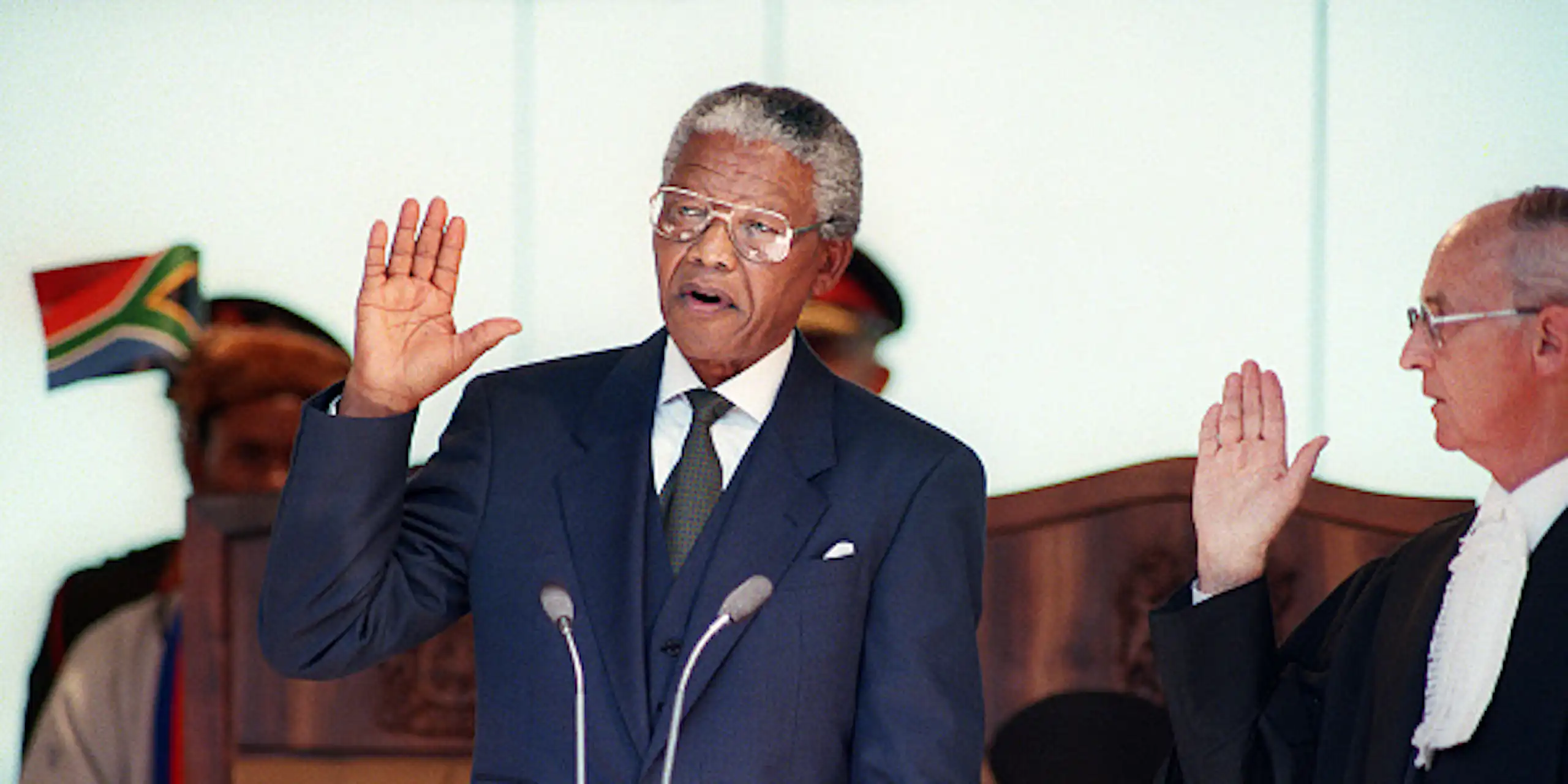 A man wearing a suit and tie raises his right hand to take an oath, wtacxhed by a male judge.