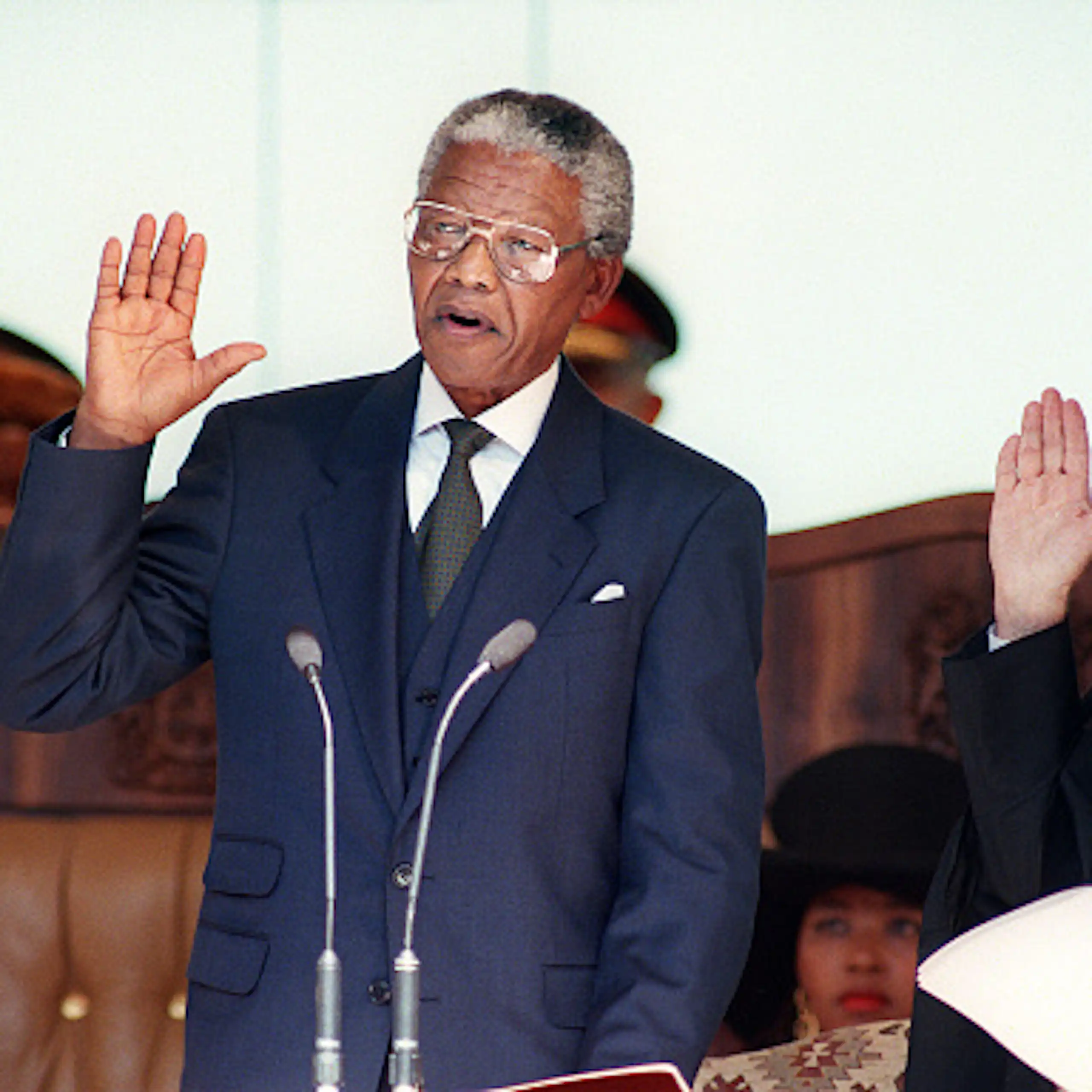 A man wearing a suit and tie raises his right hand to take an oath, wtacxhed by a male judge.
