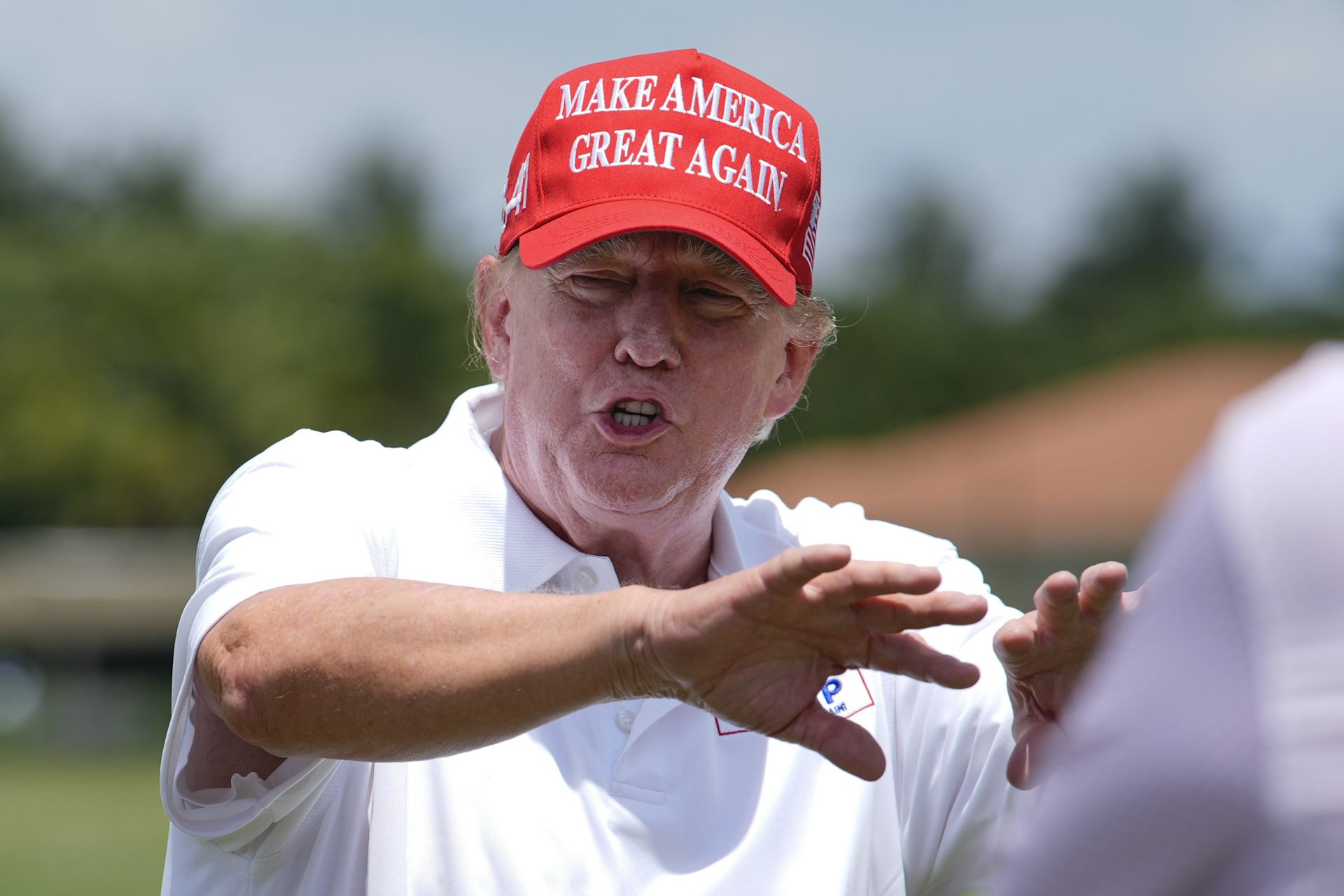 A man in a white golf shirt and a red Make America Great Again hat speaks and gestures on a golf course.