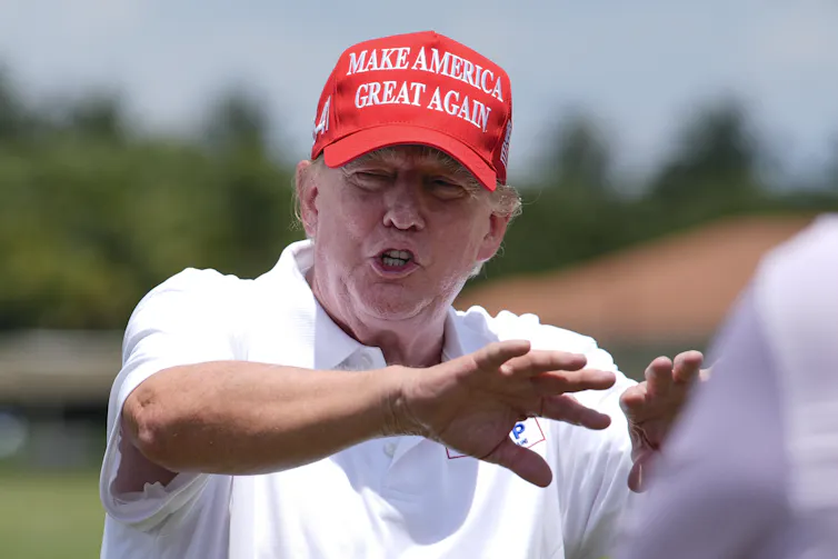A man in a white golf shirt and a red Make America Great Again hat speaks and gestures on a golf course.