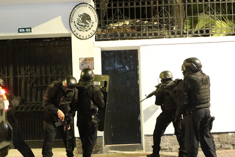 people in helmets and riot gear carrying guns stand outside a white building.
