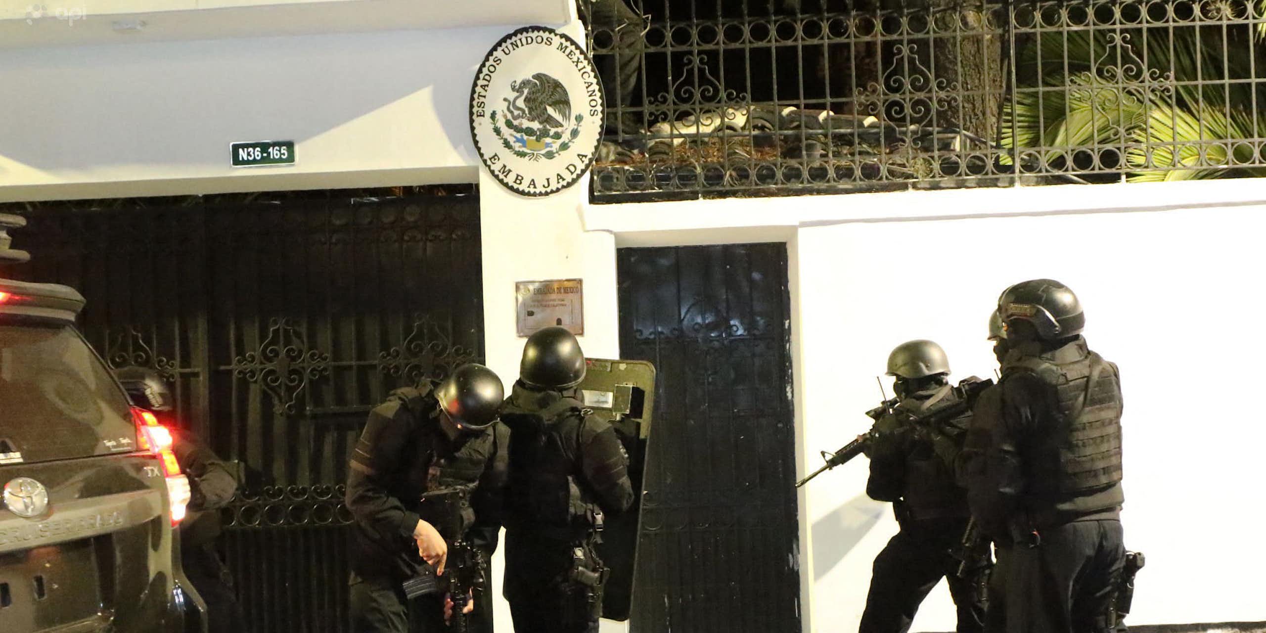 people in helmets and riot gear carrying guns stand outside a white building.