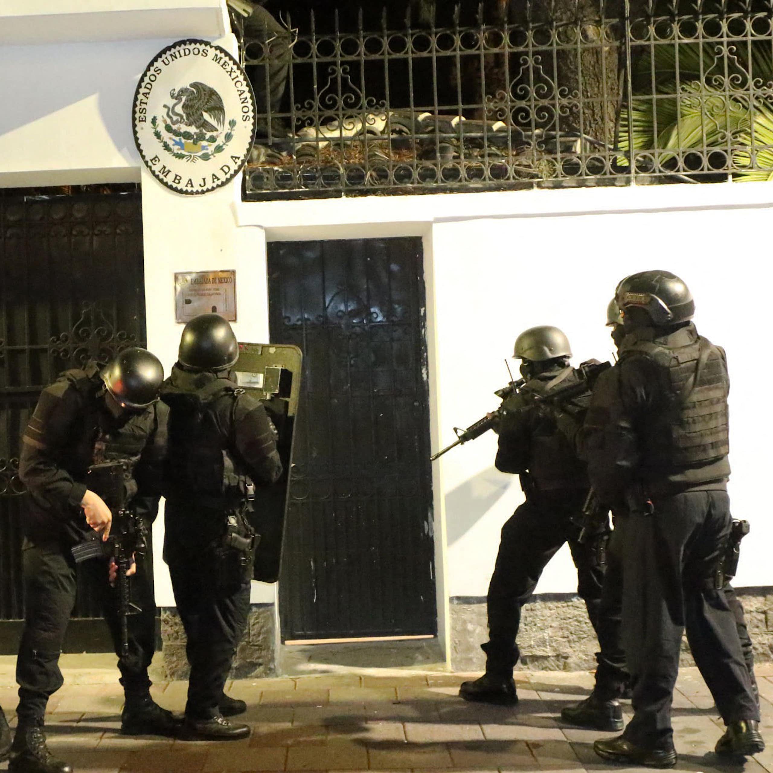 people in helmets and riot gear carrying guns stand outside a white building.