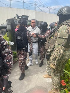 A man in handcuffs and a white jumpsuit is led away by men in fatigues.