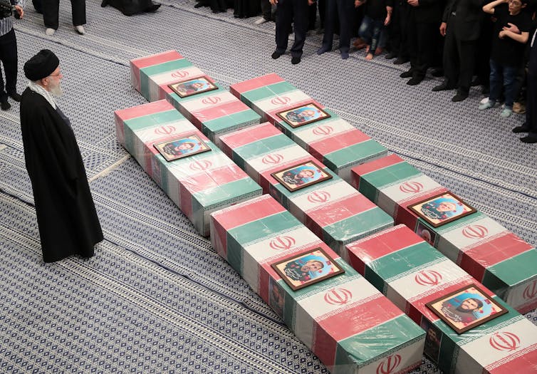 A man in traditional Iranian garb stands next to a line of coffins in red, white and green -- the colors of the Iranian flag.