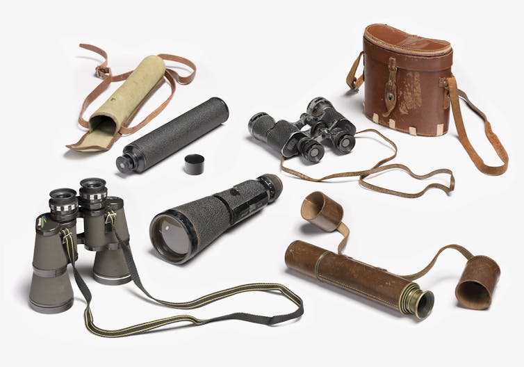 white background with six old pairs of binoculars and their brown cases laid out on display